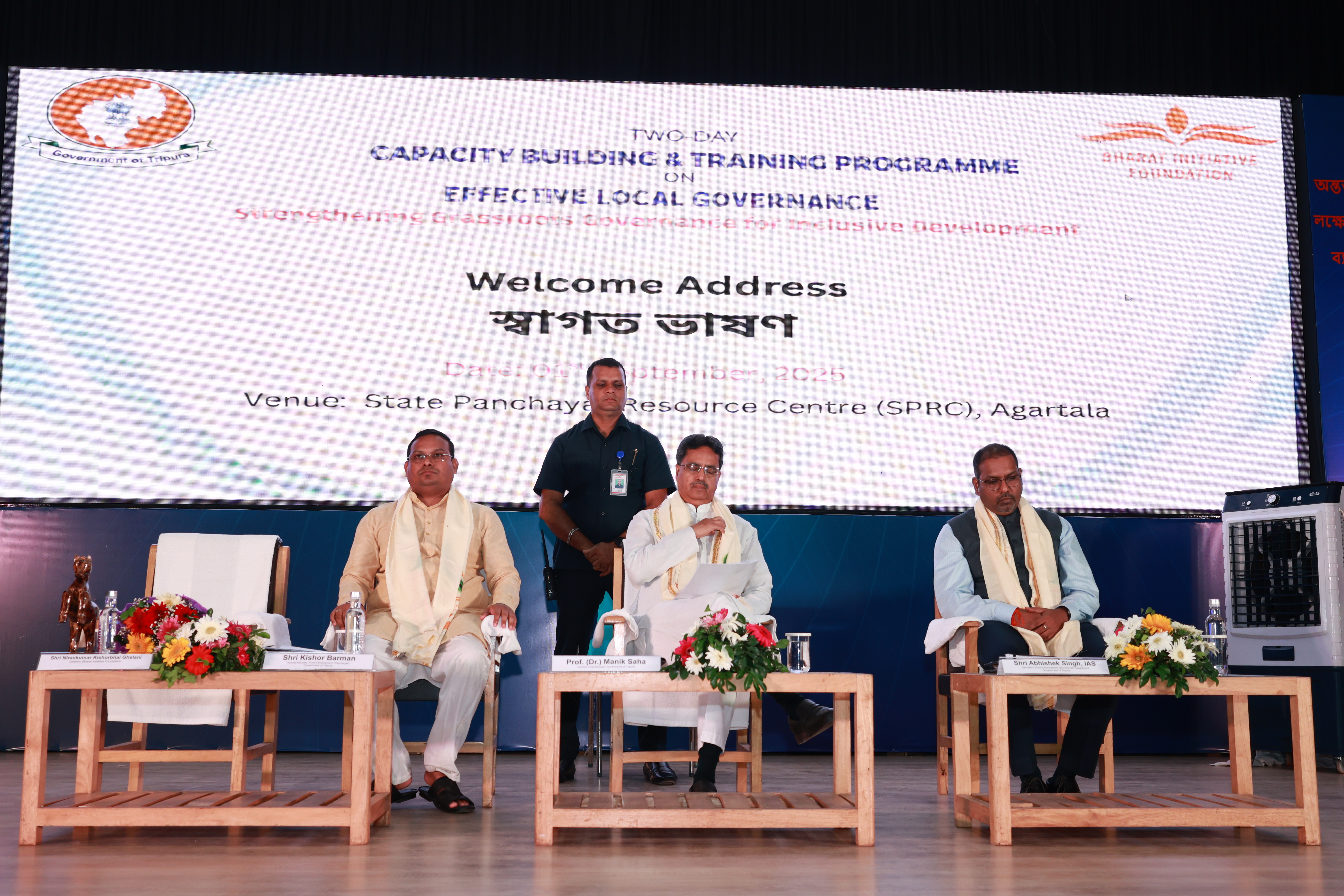 A speaker reading the welcome address on stage, with the screen showing Welcome Address and the date 1 September 2025